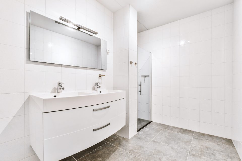 Interior of modern bathroom with rectangular mirror and clean sinks attached to white tiled wall near shower cabin in modern washroom